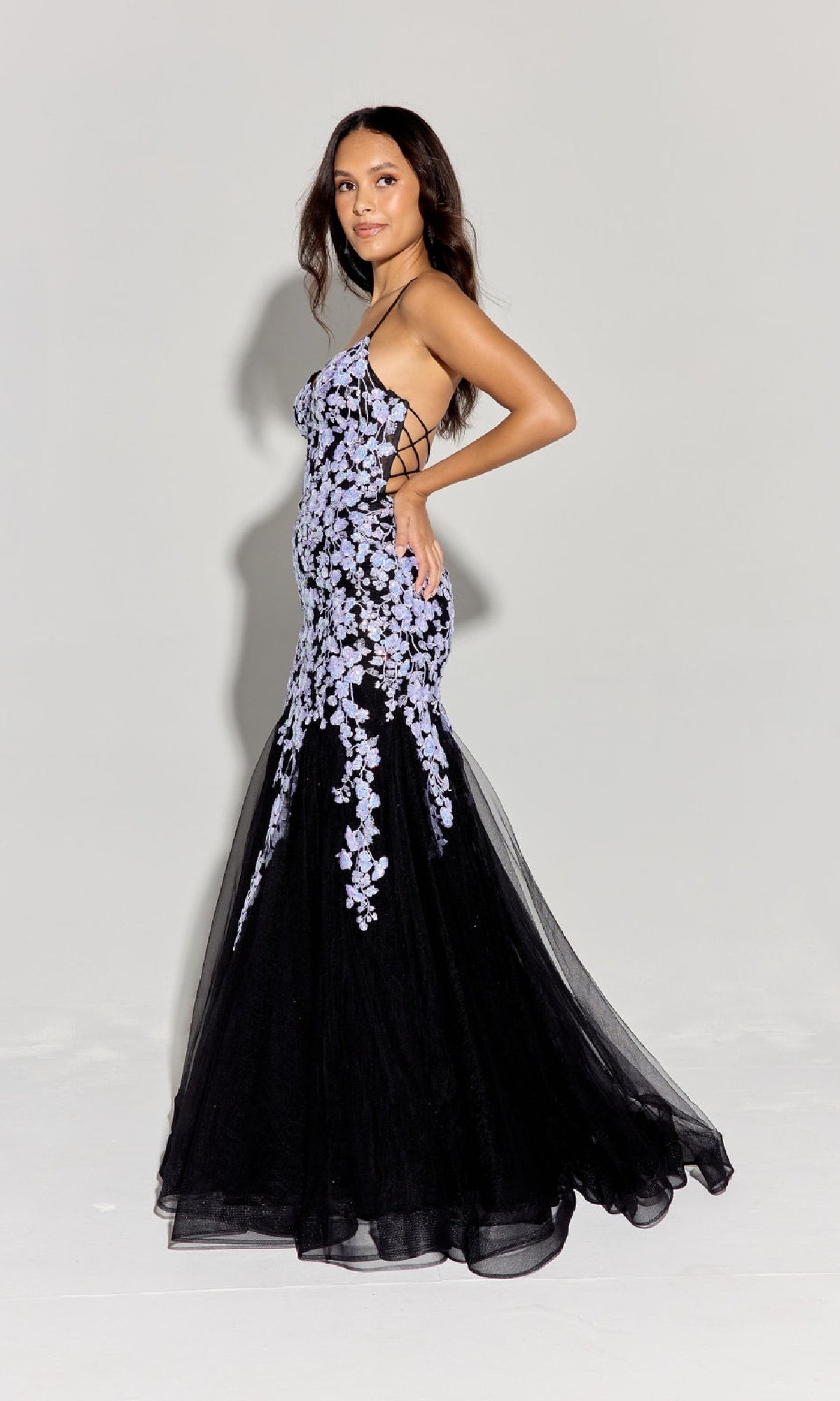 A woman stands in profile wearing the Sequin-Floral Long Mermaid Prom Dress: Eureka 9696, featuring black fabric with purple sequin floral embroidery and appliqué, set against a plain light background.